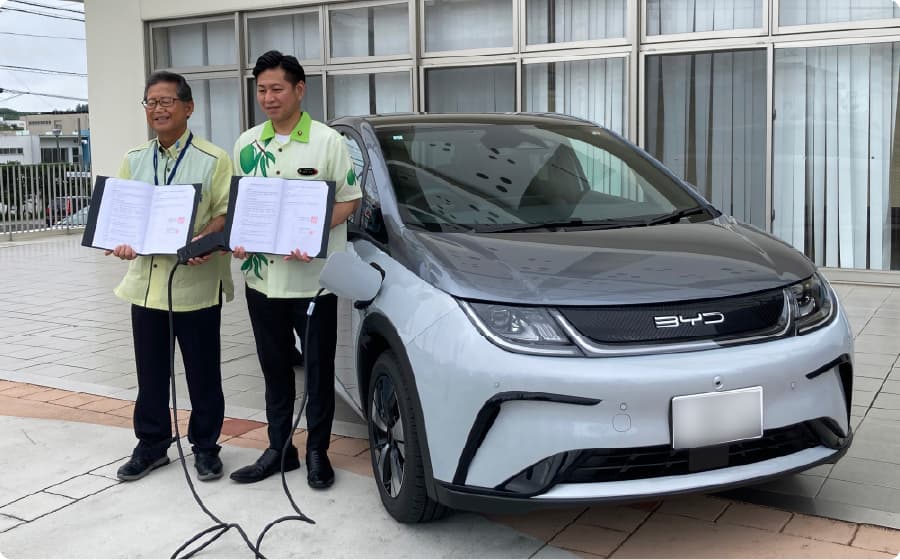 協定書を持つ二人の男性と、その横に停車している電気自動車（EV）